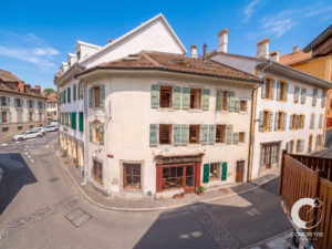 Corner white building with green shutters on a sunny European street; other pastel buildings line the block under a blue sky.