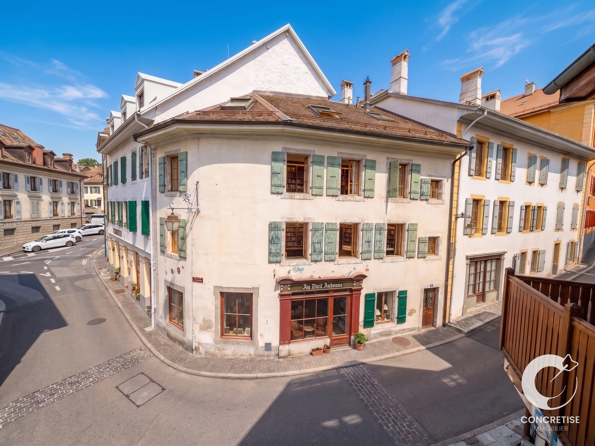 Corner white building with green shutters on a sunny European street; other pastel buildings line the block under a blue sky.