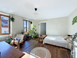Bright airy living room with many potted plants, wooden floors, and large windows letting in sunlight; a bed on the right and seating area on the left.
