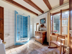 Bright rustic bathroom with a curved blue-tiled shower, white tiled walls, and exposed wooden ceiling beams near a large window with curtains.