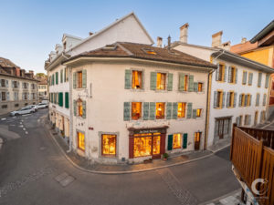 Corner building with green window shutters and warmly lit shop windows on a narrow European street at dusk/early evening.