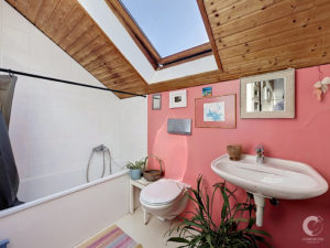 Pink-walled bathroom with a white pedestal sink, toilet and bathtub under a wooden slanted ceiling with a skylight two-panel window.