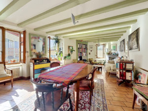 Sunlit living and dining area with pale green ceiling beams, a long wooden table covered by a plaid cloth, and bookshelves along the wall.