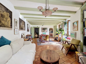 Bright living room with white sofa, round wooden coffee tables, and green-beamed ceiling; framed art on walls and bookshelf lining the right wall.