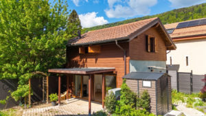 Brown wooden house with a red-tiled roof, large glass doors, and a lush green tree to the left under a bright blue sky