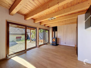 Sunlit living area with exposed wooden beams, wood panel walls, and large glass sliding doors opening to outside.