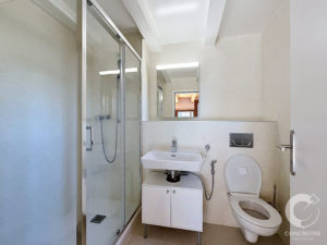 Modern bathroom with glass shower enclosure, white sink, and wall-mounted toilet in a beige-tiled room.