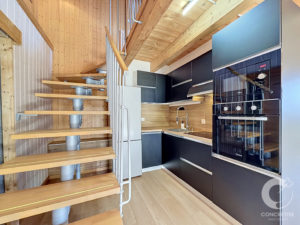 Open-plan kitchen with black high-gloss cabinets, built-in double ovens, and a wooden open-riser staircase against wood paneling and light flooring.