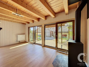 Living room with exposed wooden ceiling beams, light wood flooring, a radiator on the left wall, and large glass doors opening to an outdoor patio; a black stove pipe is on the right.