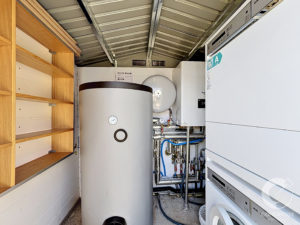 Tall vertical water heater in a utility/boiler room, with exposed pipes and a white boiler on the right and wooden shelving on the left.