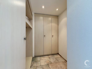 Narrow hallway closet with tall light-wood cabinets and open shelving on the left, blue textured wall on the right.