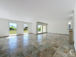 Bright, empty living area with large sliding glass doors and windows, natural light flooding a tiled floor. View of outdoors is visible through doors.
