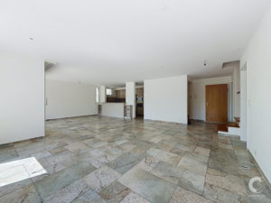 Open-plan living area with tiled floor, white walls, and a partial kitchen in the back; pendant light fixtures hanging from the ceiling.