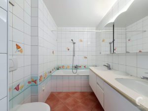 Bright bathroom with white tiled walls, a bathtub along the left, floral tile accents, and a long white countertop with two sinks and a mirror above.