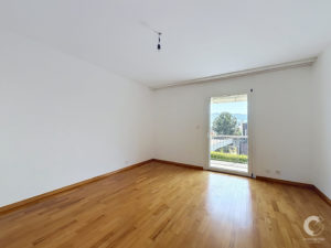 Empty bright room with light wood floors, white walls, and a single hanging light fixture; door to a balcony with a view of trees outside.