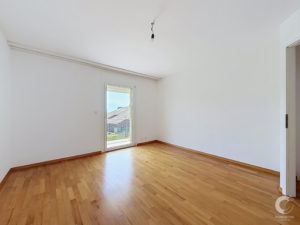 Bright empty room with white walls and warm parquet flooring, a glass door leading to a balcony, and a single hanging light bulb from the ceiling.
