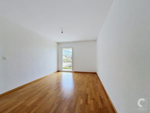 Empty rectangular room with light wooden floor, white walls, and a doorway with a window at the far end; a single hanging light bulb in the ceiling.