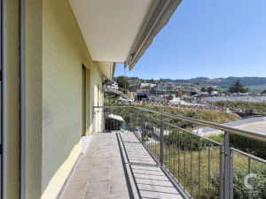 Balcony walkway along a yellow building with a metal railing and tiled floor, overlooking a construction site and distant hills under a clear blue sky.