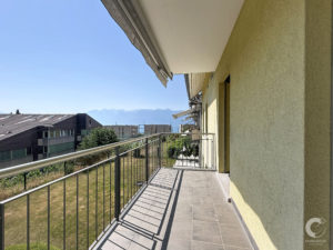 Balcony walkway with tiled grey floor and metal railing, yellow stucco wall on the right, blue sky and distant mountains in the background.