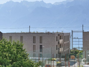 Rooftop view of a beige apartment building with a metal fence and utility lines, with hazy blue mountains in the background and green trees in the foreground.
