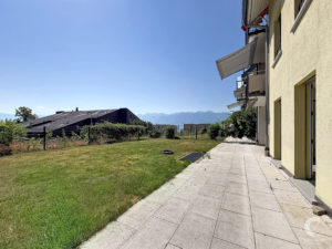 Exterior view of a beige apartment building with multiple balconies and retractable awnings along a paved patio, with a grassy lawn and distant mountains under a clear blue sky.