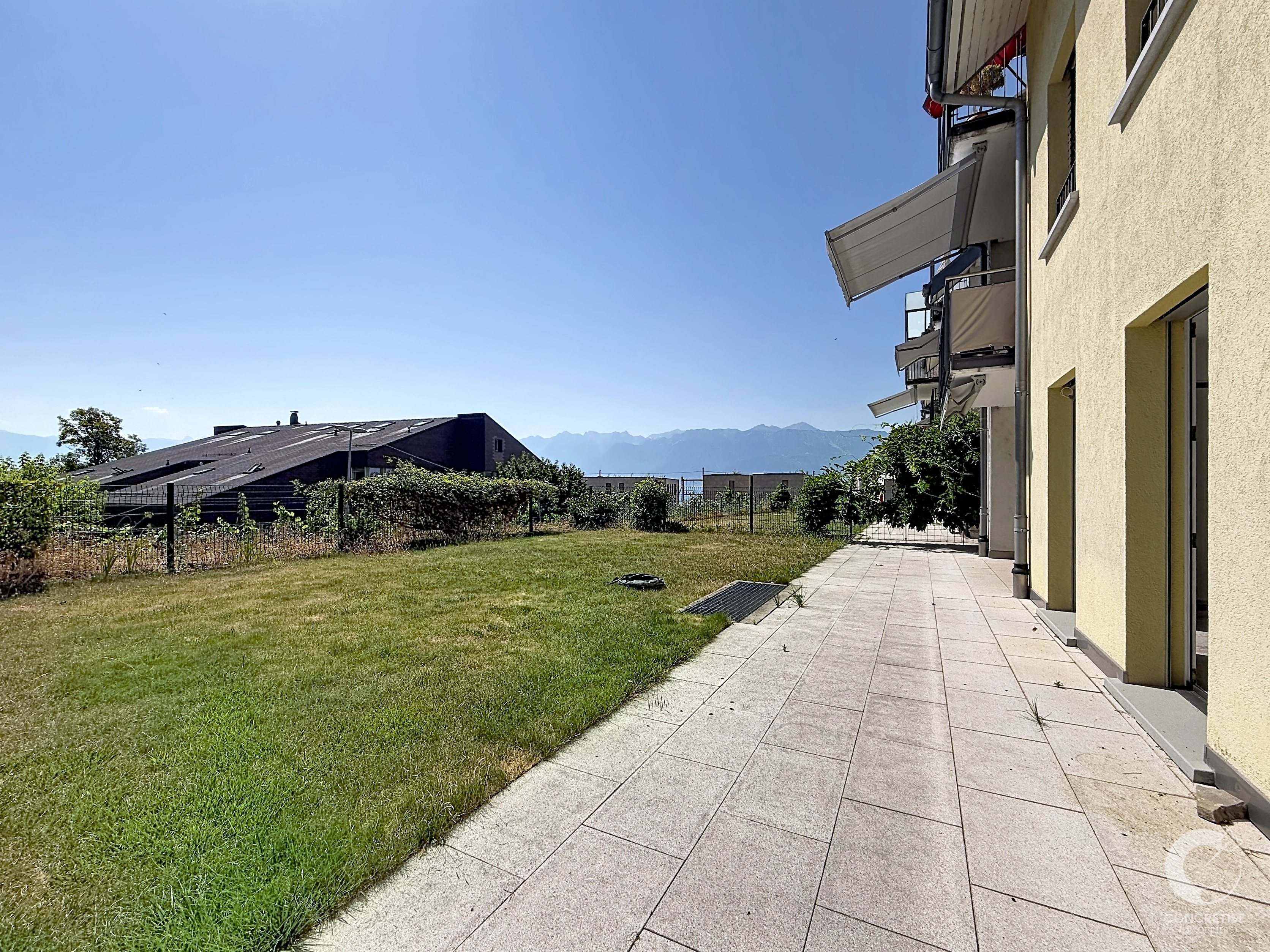 Exterior view of a beige apartment building with multiple balconies and retractable awnings along a paved patio, with a grassy lawn and distant mountains under a clear blue sky.