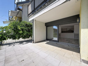 Ground-level patio with large sliding glass doors into a modern living space, beige tile floor and dense hedge along a metal fence. (Informative)