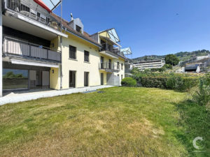 Yellow apartment building with balconies and glass canopies, a grassy lawn in front and a hillside in the distance under a blue sky.