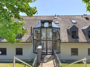 Residential building with a glass atrium and red-tiled roof, framed by green tree branches in the foreground.