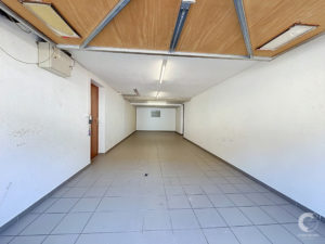 Long empty corridor with white walls, tiled floor, and fluorescent lights; wooden door on the left and exposed ceiling panels.