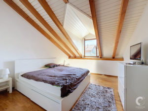 Bedroom with a white platform bed, brown duvet, and nightstand under a sloped ceiling with exposed wooden beams.