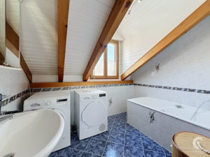 Attic bathroom with wooden beams, a window, washing machines, and a bathtub along tiled walls and blue-tiled floor