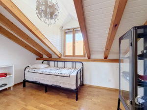 Bedroom with sloped ceiling and exposed wooden beams, metal-framed bed, window letting in light, and a glass-front cabinet on the right.