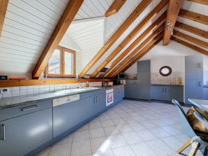 Kitchen with grey cabinets along a tiled counter under a pitched ceiling with exposed wooden beams and white paneling on the walls/ceiling in an attic space.