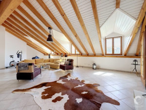 Spacious living room with exposed wooden ceiling beams, white paneled vaulted ceiling, large windows, and a brown-and-white cowhide rug on tiled floor; tan sectional sofa and chairs scattered throughout.