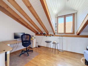 Home office in an attic with white paneled walls, exposed wooden beams, and a large window letting in daylight. A desk, monitor, and office chair sit along the wall.
