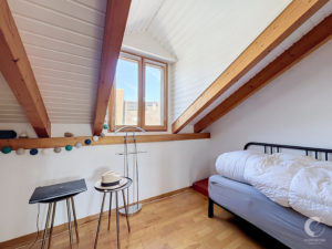 Attic bedroom with sloped white paneled ceiling, large wooden window, bed with white bedding, and colorful string lights along a shelf.