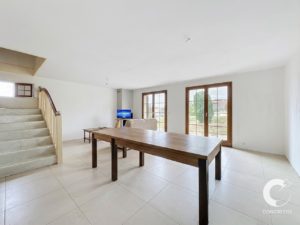Bright, empty living/dining area with a long wooden table, white tiled floor, and a staircase on the left; large glass doors open to the yard.