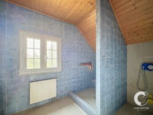 Under-construction bathroom with blue tiled walls, a wooden slanted ceiling, a white-framed window, radiator beneath, and a partially tiled shower niche.