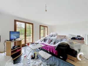 Living room under renovation: sofa and area covered in plastic, TV on a small wooden stand, scattered tools and supplies on the floor.