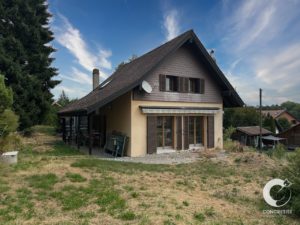 Chalet-style house with a steep roof, brown shutters, and a satellite dish, set in a dry grassy yard under a blue sky.
