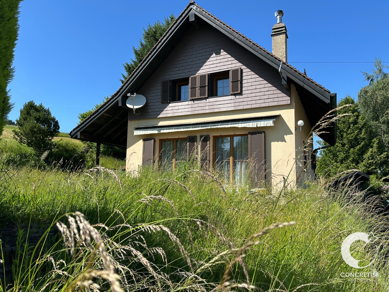 A beige house with dark brown shutters and a steep gabled roof, set in tall grass under a clear blue sky.