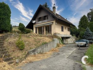 Beige house on a hillside with a gravel driveway, a covered porch, and a small trailer parked to the right.",