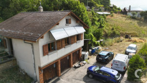 Two-story white house with brown shutters and a tiled roof, cars parked in a driveway on a hillside.