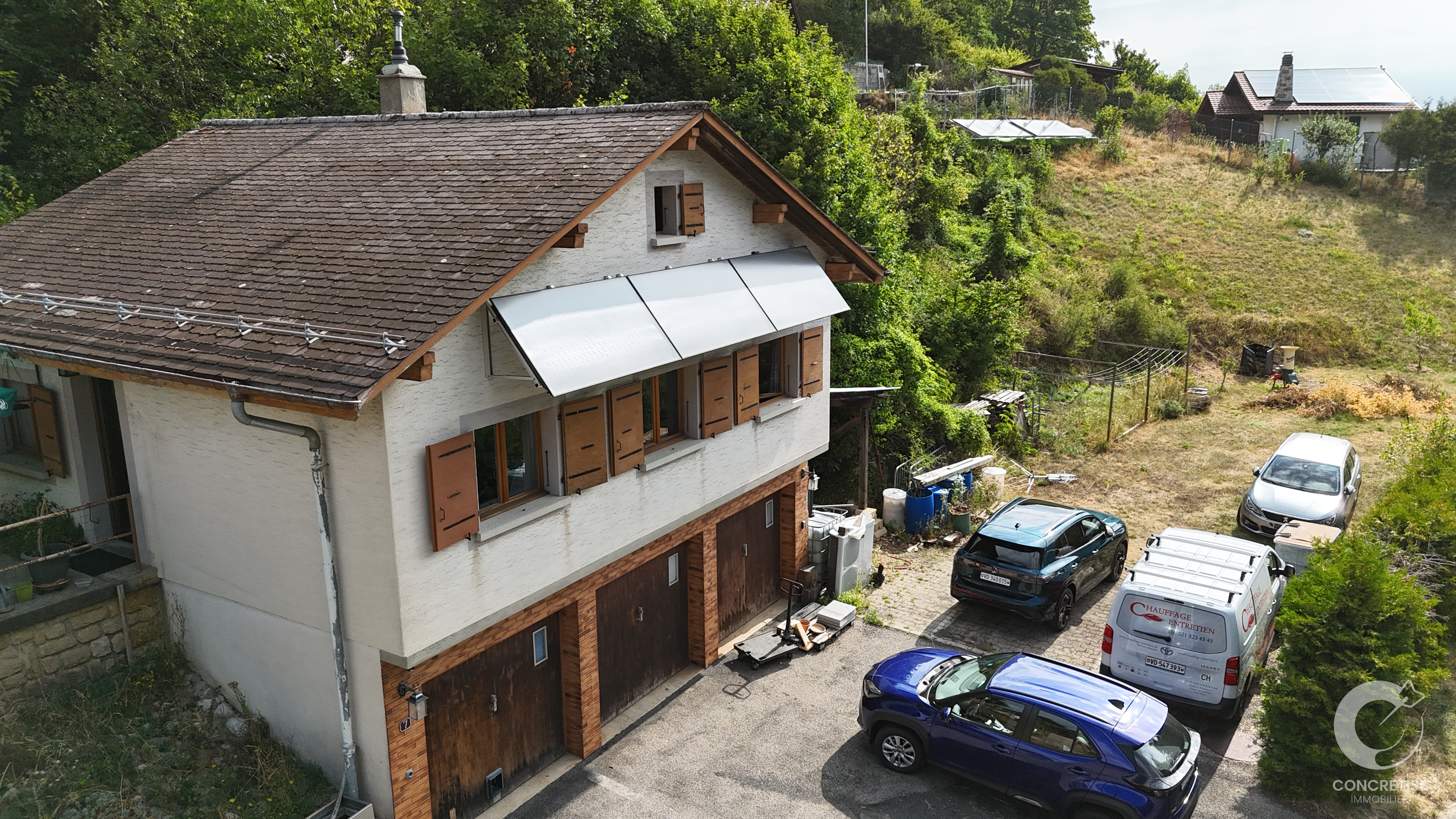 Two-story white house with brown shutters, a metal awning over windows, and a hillside yard with several parked cars nearby.