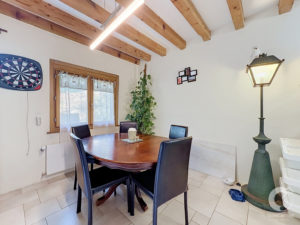 Dining area with a round wooden table and black chairs, exposed wooden ceiling beams, and a wall-mounted dartboard on the left.