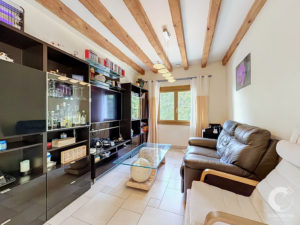 Living room: black wall unit with glass cabinets, glass coffee table, brown leather sofa, wooden ceiling beams, and a window letting in daylight.