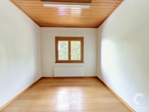 Narrow empty attic room with a wood-paneled ceiling, white walls, a small wooden-framed window at the far end, and a radiator beneath it, bathed in natural light.