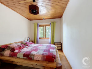Bedroom with a wooden ceiling and a large wooden bed covered in red‑pink patchwork quilt; window with lime green and lavender curtains.