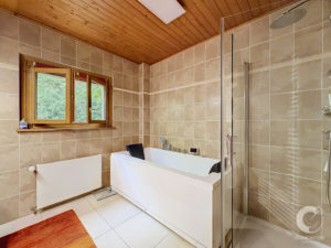 Modern bathroom with a white freestanding tub, glass shower enclosure, beige tiled walls, and a wooden ceiling near a window.